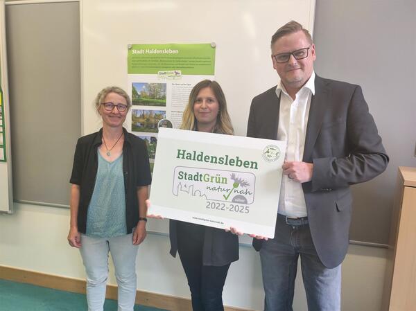 Christina Wiegmann, Abteilungsleiterin Grünanlagen, Stephanie Otto, Bauamt und Dezernent Oliver Karte mit dem Auszeichnungsschild, das die Zertifizierung bescheinigt.